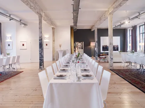 Stilvolle Eventlocation "Helmkehof" mit langen, festlich gedeckten Tischen, rustikalen S&auml;ulen und moderner Kunst an den W&auml;nden.