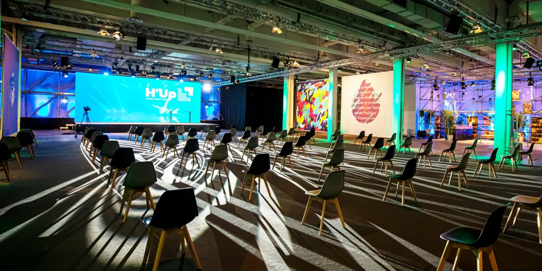 Beleuchteter Eventraum mit vereinzelten Stühlen, großer LED-Bühne mit „H’Up“-Logo und kreativer Wandgestaltung.
