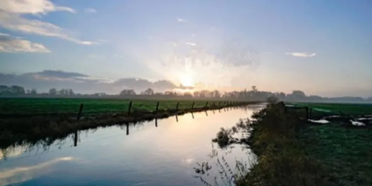 Sonnenaufgang über einer weiten Wiesenlandschaft mit einem ruhigen Wassergraben, der den Himmel reflektiert.