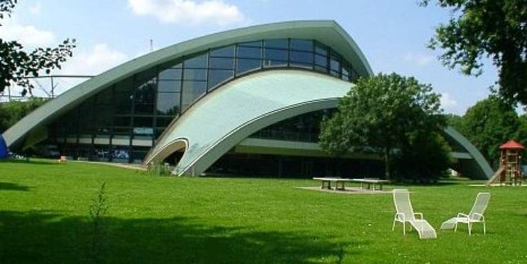 Schwimmhalle, davor grüne Wiese mit zwei weißen Liegestühlen