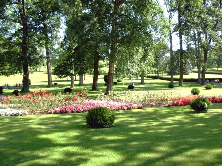 Eine große Rasenfläche ist von Blumenbeeten und und wenigen Büschen unterbrochen, dahinter stehen Bäume.