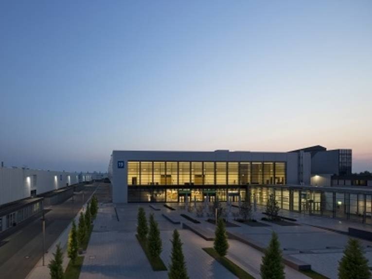 Moderne Messehalle mit großer Glasfassade, beleuchtet bei Abenddämmerung, umgeben von einem gepflasterten Platz mit Bäumen.