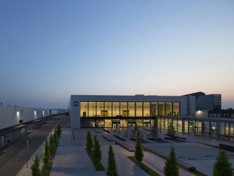 Moderne Messehalle mit großer Glasfassade, beleuchtet bei Abenddämmerung, umgeben von einem gepflasterten Platz mit Bäumen.