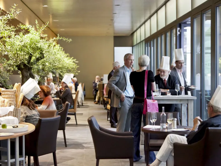 Elegantes Restaurant "Schlossküche Herrenhausen" mit großen Olivenbäumen, Gästen in Kochmützen und lockerer Gesprächsatmosphäre.