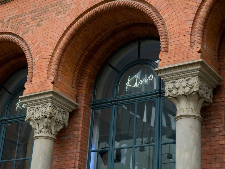 Eine stuckverzierte Häuserfassade mit Fenstern und der Leuchtschrift Kino
