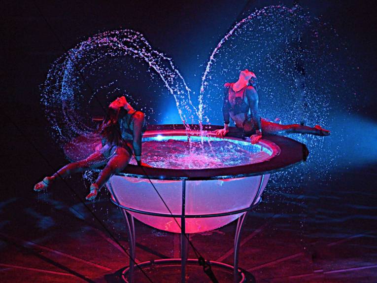 Zwei Frauen in akrobatischer Pose an einem durchsichtigen Wasserbecken auf. Wassertropfen in der Luft.